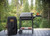 PK Grills PK300 Start Cookin' Bundle - Graphite, a portable charcoal grill with a side tray, a black carrying bag with the PK logo, set on a wooden deck outdoors surrounded by green trees.