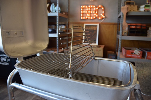PK Grills Stainless Steel Cooking Grid for Original PK, a stainless steel cooking grate inside a portable grill with a background of a store with a BBQ sign.