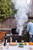 PK Grills The PK360 Grill & Smoker - Classic Silver, outdoor barbecue grill with smoke rising, set in backyard with food and drinks on table.